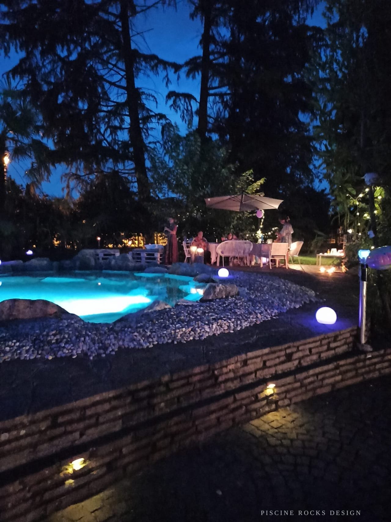 Piscina Rocks Design illuminata di sera, con gli ospiti di un evento ai tavoli sullo sfondo