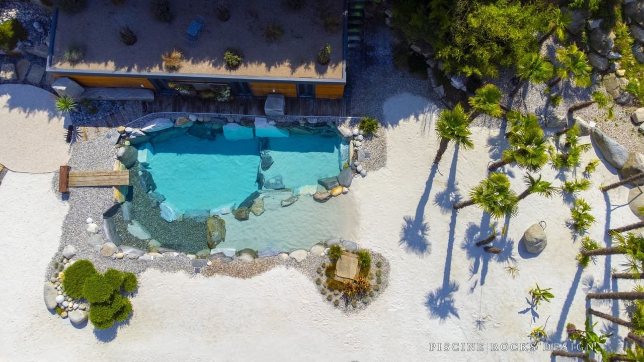 Vista aerea di una piscina Piscine Rocks Design con spiaggia in sabbia bianca, palme e massi monolitici